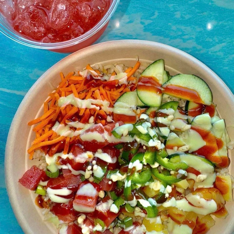 Loaded rice bowl topped with vegetables, watermelon, carrots, cucumber, peppers, creamy dressing, and red sauce beside an iced red drink.