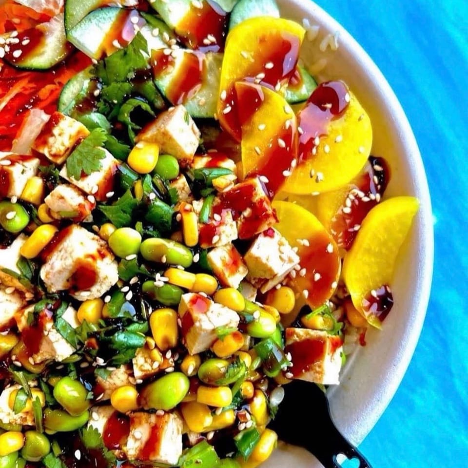 Colorful salad with tofu, corn, edamame, cucumber, tomatoes, herbs, sesame seeds, and red dressing in a white bowl.