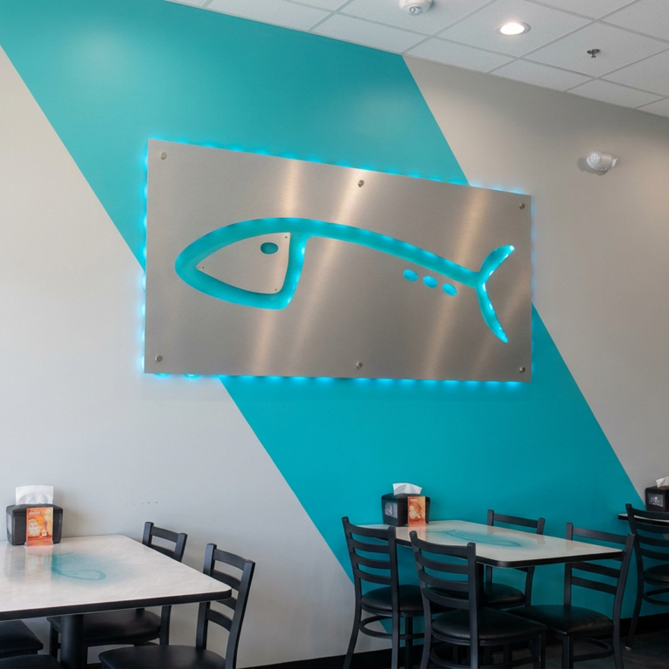 Bright seafood restaurant dining area with black tables and chairs, turquoise accent wall, and illuminated fish-shaped metal sign.