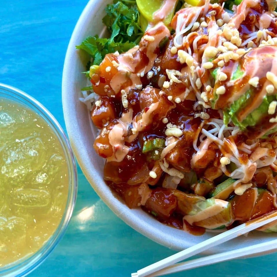 Loaded poke bowl with glazed salmon, avocado, greens, lime, crunchy toppings, and creamy sauce beside a lime drink and chopsticks.