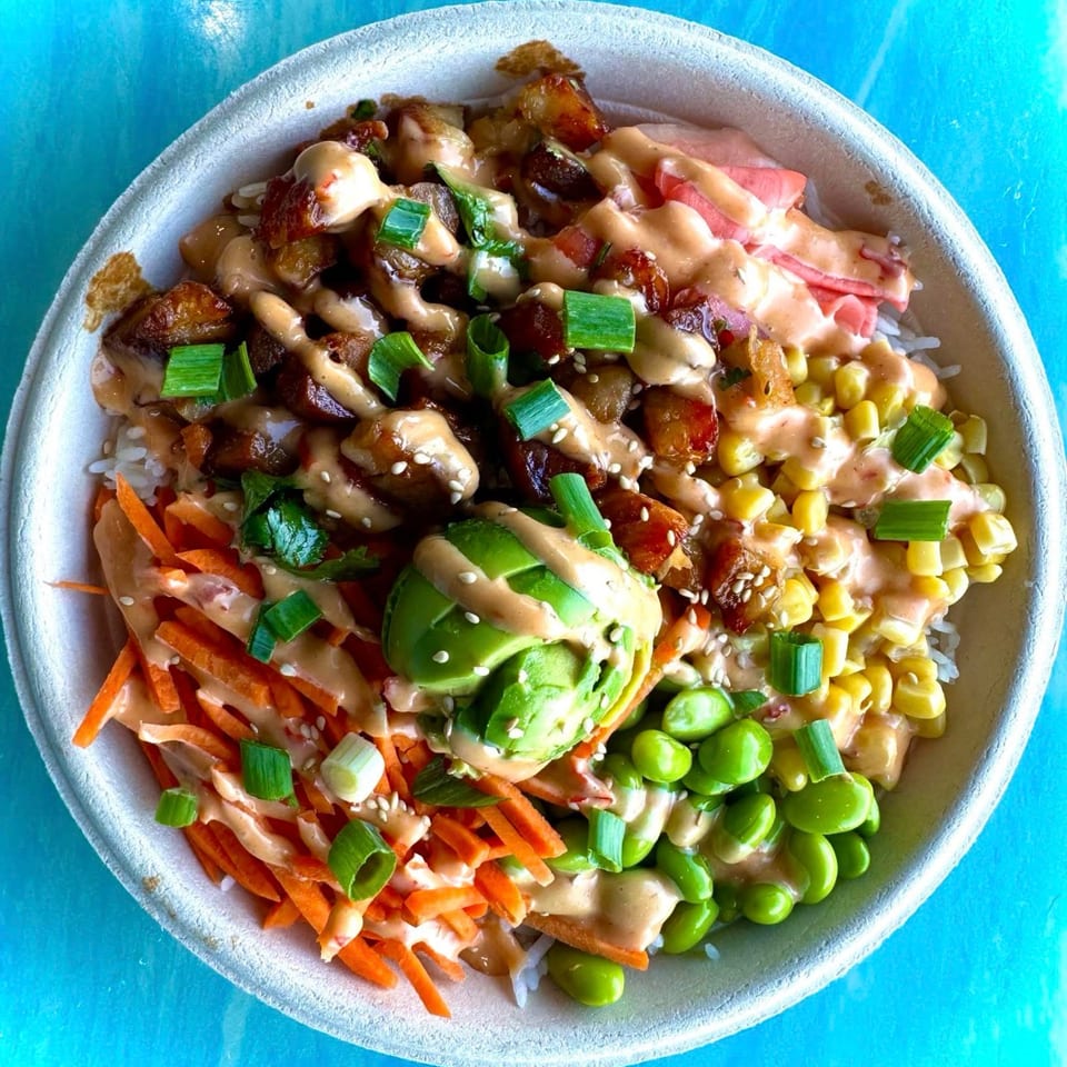 Colorful rice bowl topped with roasted vegetables, avocado, edamame, corn, carrots, pickled ginger, green onions, sesame seeds, and creamy sauce.