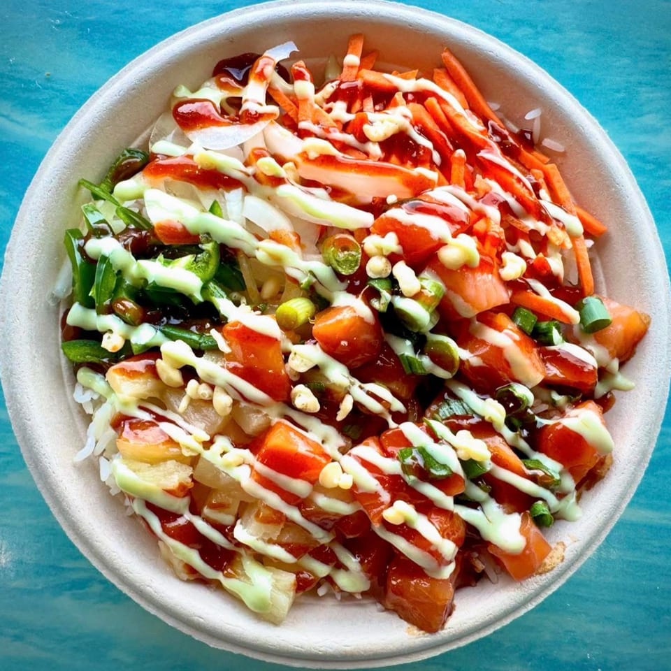 Bowl of rice topped with vegetables, carrots, green onions, corn, and tomatoes, drizzled with red and creamy sauces.